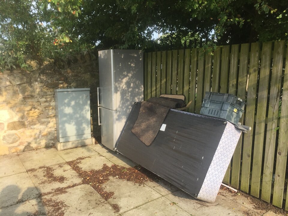 A discarded fridge freezer and mattress left out for collection beside a garden fence